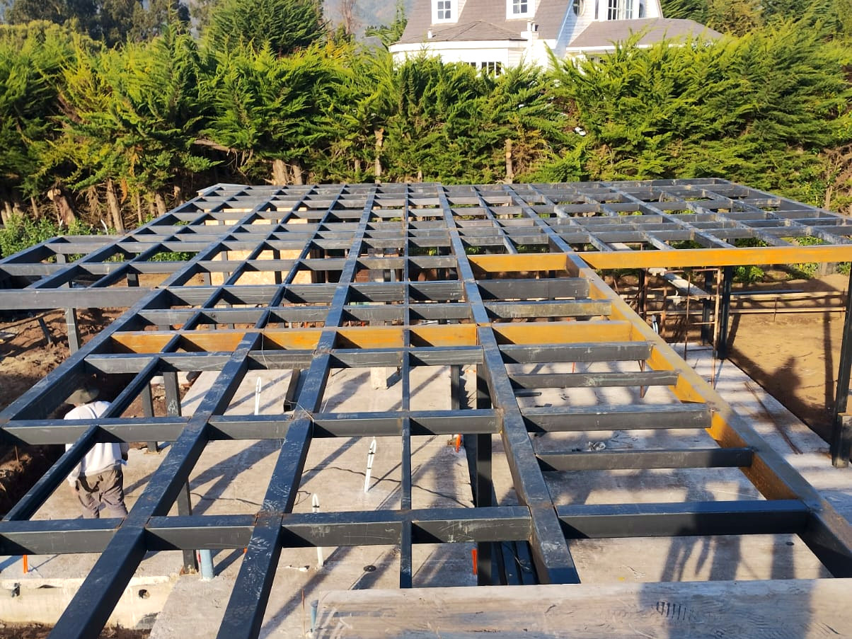 Estructura metálica en construcción, con una vivienda al fondo